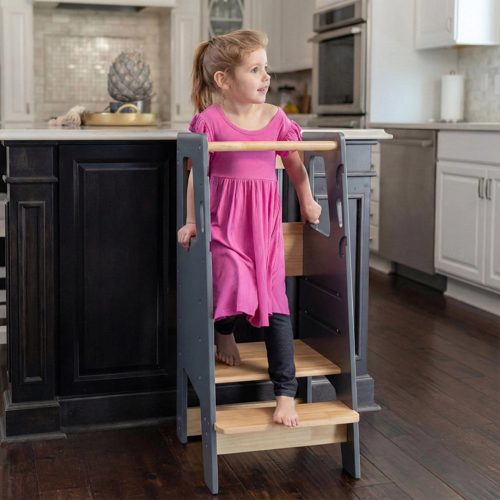 Front angled view of a toddler girl standing on a grey solid rubberwood Montessori stool by a dark island