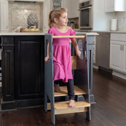 Front angled view of a toddler girl standing on a grey solid rubberwood Montessori stool by a dark island