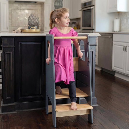 Front angled view of a toddler girl standing on a grey solid rubberwood Montessori stool by a dark island