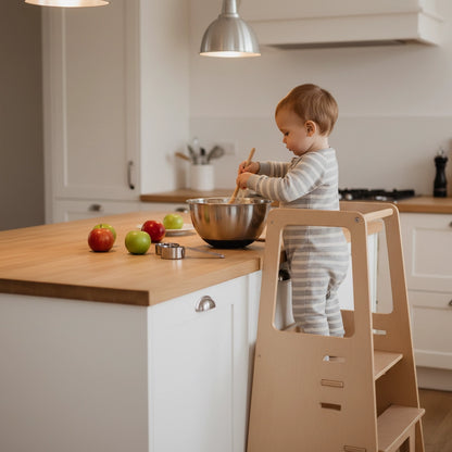 A toddler stands on the birch plywood Montessori standing stool while mixing ingredients in a metal bowl
