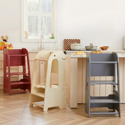 Three toddler helper towers in red, natural wood, and gray finishes lined up against a kitchen island