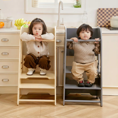 Two children sitting inside natural wood and gray step stool towers facing forward in a kitchen