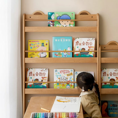 A child drawing at a wooden desk with a front-facing Montessori book rack filled with books in the background