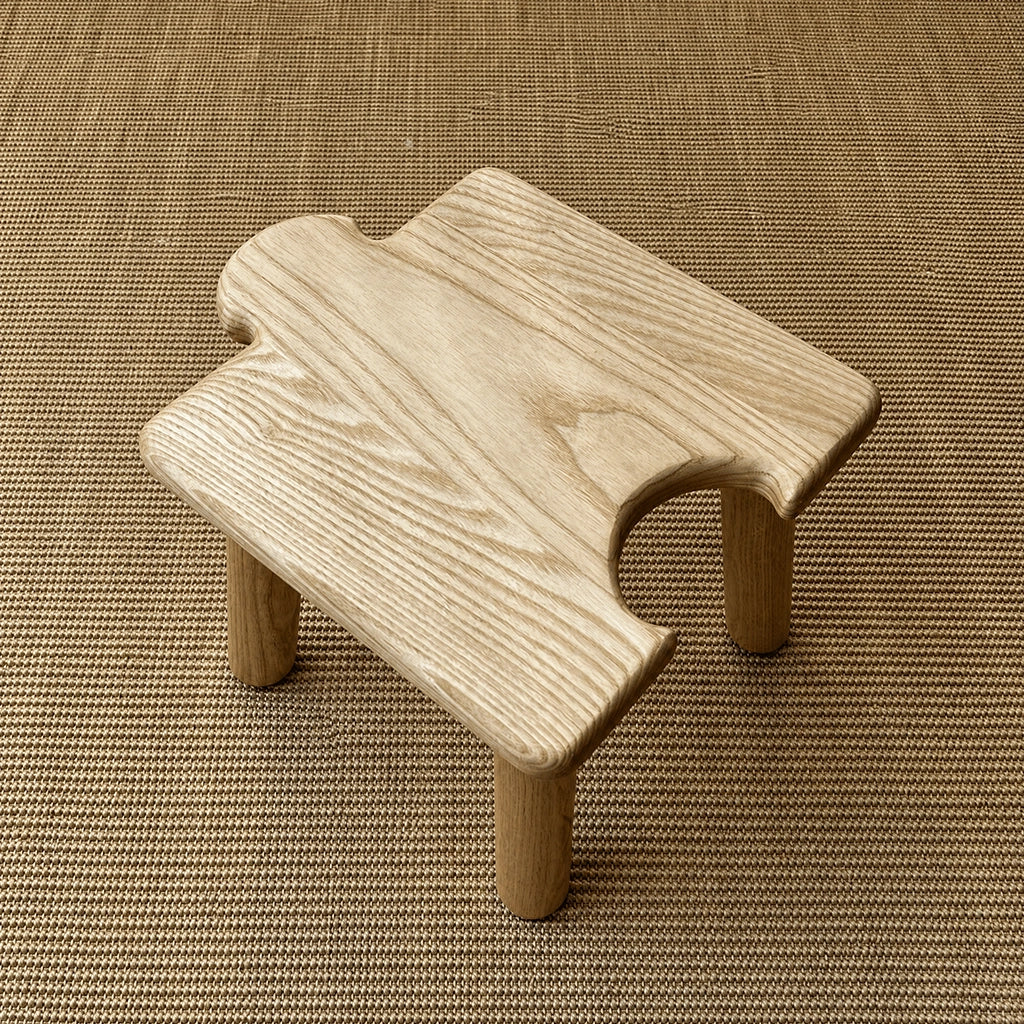 High-angle view of a light solid wood interlocking puzzle stool with a rear-facing cutout on a woven mat