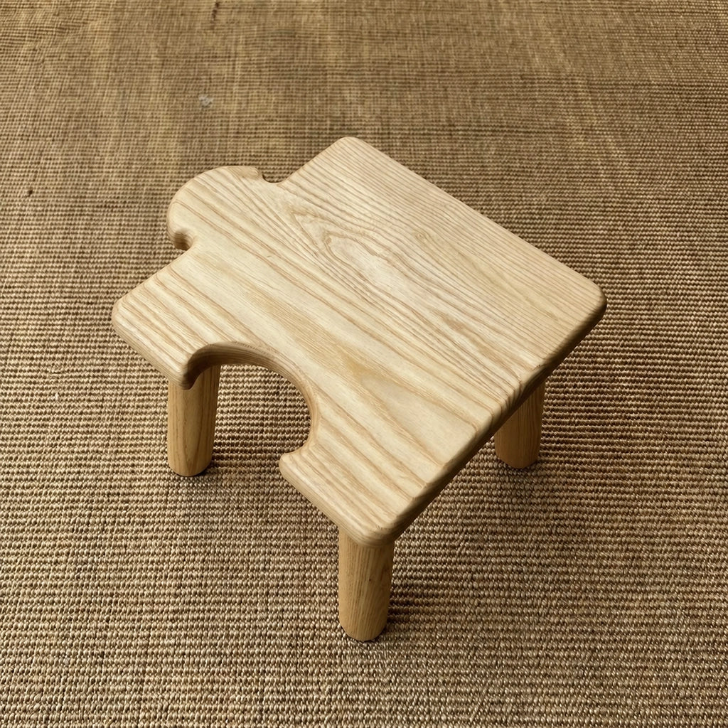 Top-angled view of a light solid wood kids puzzle stool with a side interlocking cutout on a woven rug