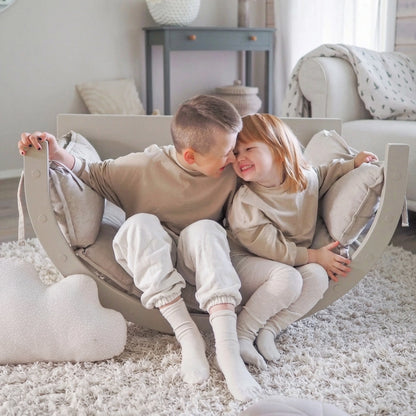 Two smiling children sitting inside the padded wooden Montessori rocker on a fluffy white rug