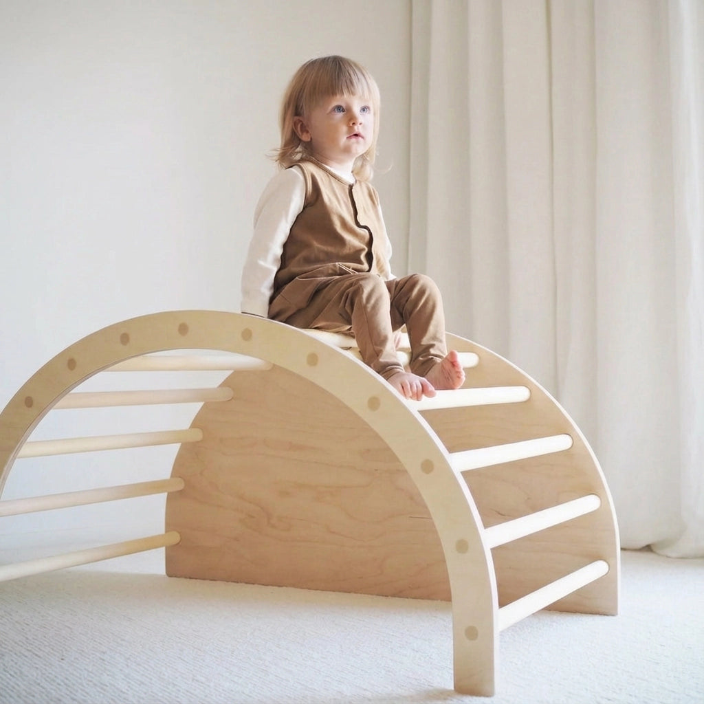 A toddler sitting on top of an upside-down natural wood Montessori climbing arch ladder