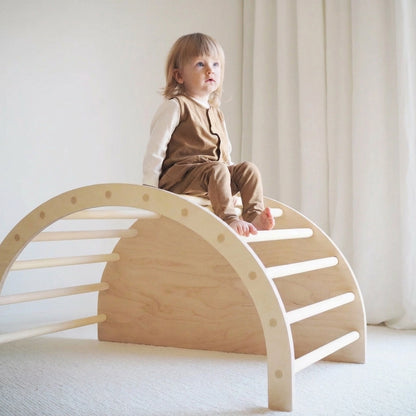 A toddler sitting on top of an upside-down natural wood Montessori climbing arch ladder